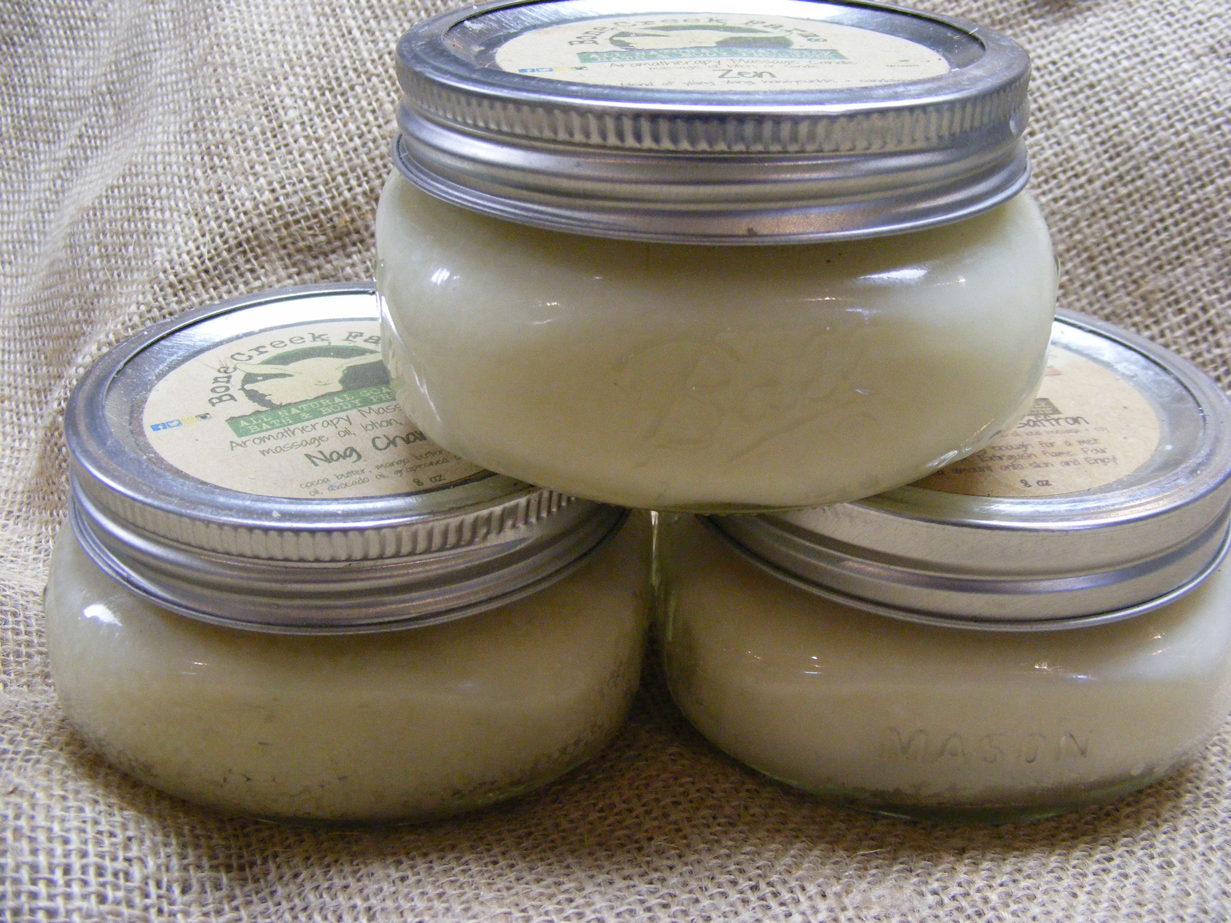 Tisha+Carr+-+candles Three jars of aromatherapy massage balm stacked on a burlap background, featuring labels that highlight their natural ingredients and soothing properties.