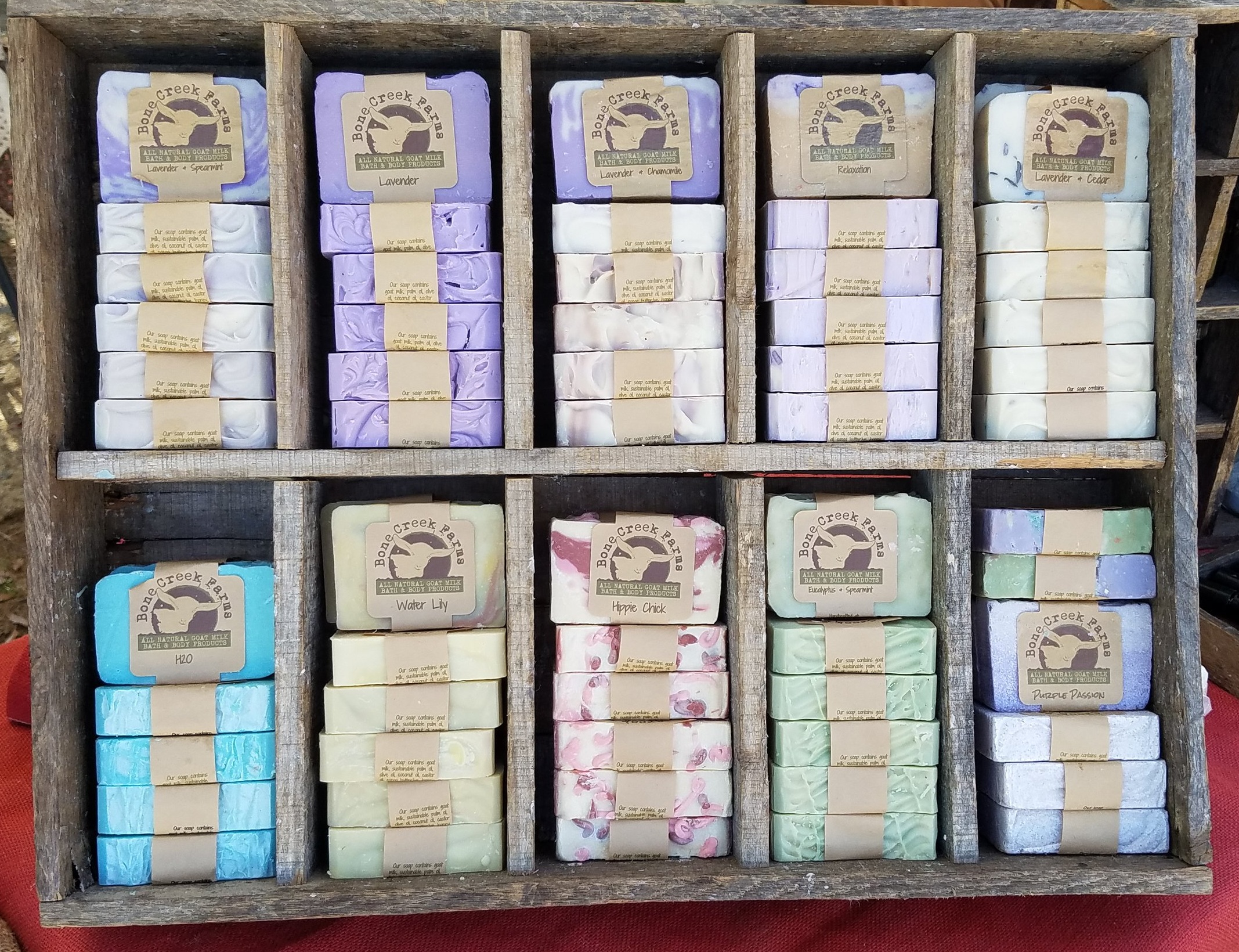 image-asset - 2026-07-24T134847.437 Artisan soap display featuring a variety of handcrafted soaps in different colors and scents, including Lavender, Water Lily, and H2O, arranged neatly in a rustic wooden crate. Each soap is labeled with its name and brand, Bone Creek Farms, highlighting the natural ingredients used.